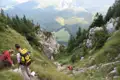 Wanderer auf einem steinigen Bergpfad. Im Hintergrund erstreckt sich ein Tal mit Wald und Wiesen.