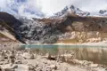 Türkisfarbener Bergsee inmitten einer Hochgebirgslandschaft mit schneebedeckten Gipfeln und Steinmännchen am Ufer.