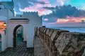 Weißes Gebäude mit Torbogen und Steinmauer am Meer bei Sonnenuntergang.