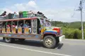 Bunter Bus mit Passagieren und Gepäck auf dem Weg durch eine ländliche Gegend.