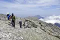 Wanderer auf einem felsigen Bergpfad mit Blick auf die umliegenden Berge und Wolken.