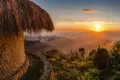 Sonnenuntergang über einer Bergkette, betrachtet von einer traditionellen Hütte mit Strohdach aus.