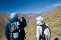 Zwei Wanderer mit Rucksäcken und Kopftüchern stehen in einer bergigen Landschaft und schauen in ein Tal.