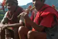 Zwei Personen in traditioneller Kleidung, möglicherweise Angehörige einer indigenen Gruppe, sitzen in einer bergigen Landschaft. Eine Person spielt auf einer traditionellen Flöte.