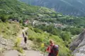 Wanderer mit Rucksäcken und Wanderstöcken auf einem Bergpfad. Im Hintergrund liegt ein Dorf in einem grünen Tal.