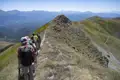 Wanderer mit Rucksäcken und Wanderstöcken auf einem schmalen, grasbewachsenen Berggrat. Im Hintergrund sind weitere Berge und Täler zu sehen.