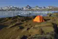 Campingplatz mit Zelt am Ufer eines Fjords in arktischer Umgebung, mit Blick auf schneebedeckte Berge und Eisberge.