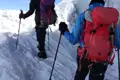 Zwei Personen beim Bergsteigen im Schnee, ausgerüstet mit Rucksäcken und Wanderstöcken. Ein Fixseil dient zur Sicherung.