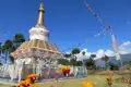 Weißer Stupa mit goldenen Verzierungen und bunten Gebetsfahnen, umgeben von einem blauen Zaun und orangen Blumen. Im Hintergrund Bäume und Berge.