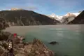 Wanderer am Ufer eines Gletschersees mit Bergen und Gletschern im Hintergrund.