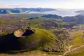 Luftbild eines Vulkankraters mit umliegender Vegetation, einem See im Hintergrund und Anzeichen von vulkanischer Aktivität in der Landschaft.