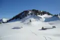 Zwei Hütten in einer verschneiten Berglandschaft mit einem bewaldeten Hügel im Hintergrund.