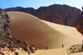 Zwei Personen in der Wüste vor einer großen Sanddüne und Felsformationen.
