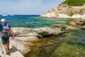 Wanderung entlang einer felsigen Küste mit türkisfarbenem Wasser und Segelbooten im Hintergrund.