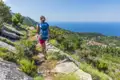 Wanderin mit Rucksack auf einem steinigen Küstenpfad. Im Hintergrund das blaue Meer und ein Dorf.