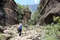 Wanderer auf einem steinigen Weg in einer Schlucht, umgeben von hohen Felswänden und Vegetation.