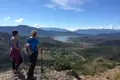 Zwei Wanderer mit Wanderstöcken pausieren auf einem Felsvorsprung und genießen den Ausblick auf das Tal, den See und das Dorf in der Ferne.