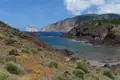 Sicht auf eine felsige Küstenlandschaft mit rötlichen Felsen und spärlicher Vegetation. Im Hintergrund sind zwei markante Felseninseln im Meer sichtbar.