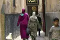 Zwei Frauen in traditionellen Gewändern gehen durch eine enge Gasse. Im Hintergrund sind Kinder und ein verzierter Türrahmen zu sehen.