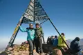 Zwei Wanderer am Gipfelkreuz, welches mit verschiedenen Namen und Botschaften beschriftet ist. Im Hintergrund sind weitere Wanderer und die Berglandschaft zu sehen.