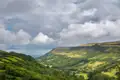 Panoramablick auf ein Tal mit üppiger Vegetation und bewaldeten Hängen unter einem bewölkten Himmel.