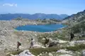 Wanderer mit Rucksäcken steigen einen felsigen Pfad hinab in Richtung eines Bergsees. Im Hintergrund erstreckt sich eine Bergkette.
