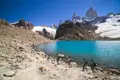 Wanderer am Ufer eines türkisfarbenen Bergsees mit Blick auf einen schneebedeckten Gipfel.