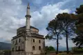 Alte Steinmoschee mit Minarett, umgeben von Bäumen und mit Bergen im Hintergrund.