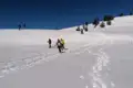 Schneeschuhwanderung in den Bergen im Winter. Mehrere Personen mit Schneeschuhen steigen einen verschneiten Hang hinauf, während im Hintergrund andere Ski fahren.