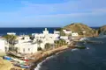 Blick auf einen Küstenort mit weißen Häusern und Fischerbooten am Meer. Im Hintergrund ist eine kleine Insel zu sehen.