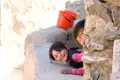 Zwei Kinder, die sich hinter einer Mauer verstecken, lächeln in die Kamera.  Zwei rote Eimer stehen auf der Mauer.