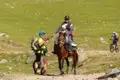 Reisende begegnen sich auf einem Weg: Ein Mann mit Kind auf dem Pferd und ein Radfahrer halten an, um miteinander zu sprechen. Weitere Radfahrer sind im Hintergrund zu sehen.