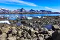 Felsiger Strand mit Eisbergen und schneebedeckten Bergen im Hintergrund.