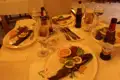 Gegrillter Fisch auf Tellern, angerichtet mit Zitronen, Orangen und Kräutern. Getränke wie Bier, Wasser und möglicherweise Wein begleiten das Essen.