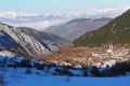 Blick auf ein verschneites Bergdorf im Tal, umgeben von schneebedeckten Bergen und Wäldern im Winter.