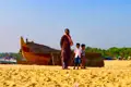 Ein Mann und zwei Kinder stehen am Strand neben einem traditionellen Fischerboot, das möglicherweise für Fischfang oder Bootsfahrten genutzt wird.
