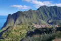Panoramablick auf ein Dorf an der Küste, umgeben von grünen Bergen und dem Meer im Hintergrund.