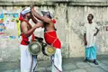 Zwei Musiker mit traditionellen Trommeln und roter und weißer Kleidung interagieren miteinander, während ein Zuschauer in blau-weißer Kleidung zusieht.
