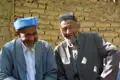 Zwei lächelnde Männer in traditioneller Kleidung vor einer Lehmziegelwand.