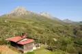 Eine einfache Berghütte mit rotem Dach inmitten einer grünen Hügellandschaft mit hohen Bergen im Hintergrund.