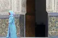 Frau in traditioneller Kleidung vor verzierter Wand mit Mosaikfliesen.
