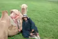 Reisender in traditioneller Kleidung pausiert neben einem Kamel auf einer grünen Wiese.