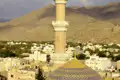 Blick auf die goldene Kuppel und das Minarett einer Moschee, eingebettet in eine Ortschaft mit Palmen und Bergen im Hintergrund.