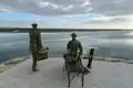 Zwei Statuen am Ufer eines Gewässers. Die stehende Figur trägt einen Koffer, die sitzende Figur ruht auf einer Bank. Beide blicken in die Ferne über das Wasser.