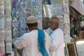 Zwei Männer begutachten traditionelle Kopfbedeckungen an einem Marktstand. Die Hüte sind in Plastik verpackt und zeigen verschiedene Muster und Farben.