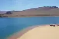 Blauer See mit Sandstrand und Personengruppe am Ufer, umgeben von kargen Hügeln.