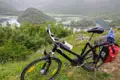 Fahrrad mit Gepäcktaschen auf einer Anhöhe, im Hintergrund ein Fluss und bewaldete Hügel.