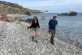 Zwei Personen wandern an einem Kiesstrand entlang, das Meer und die Küste im Hintergrund.
