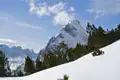 Schneebedeckter Berggipfel mit Bäumen im Vordergrund und weiteren Bergen im Hintergrund.