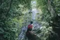 Reisender pausiert auf einem Baumstamm und betrachtet einen Wasserfall im Dschungel.
